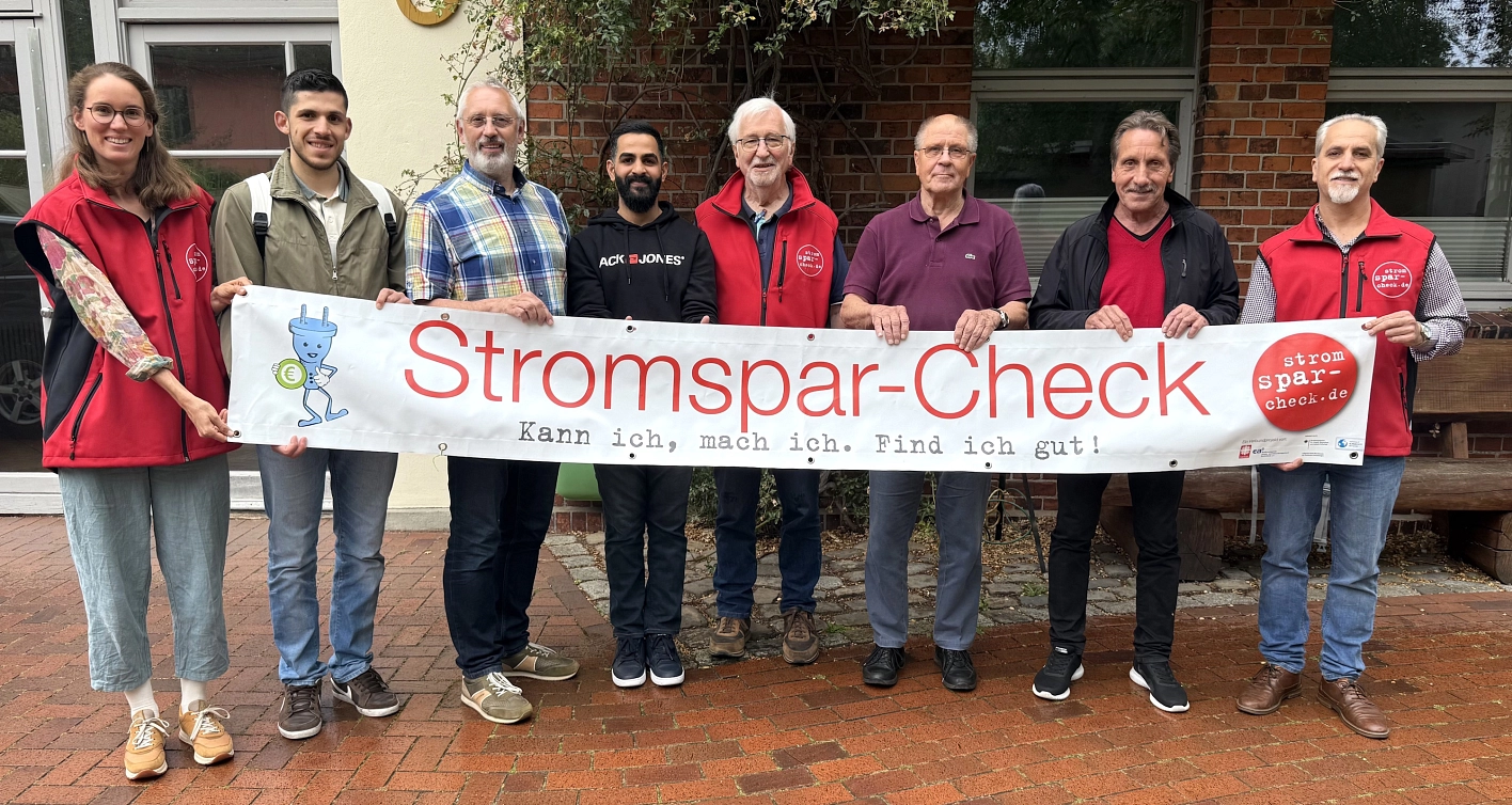 Acht Beraterinnen und Berater des Stromspar-Checks stehen aufgereiht nebeneinander und halten gemeinsam ein Transparent mit der Aufschrift "Stromspar-Check" in den Händen.